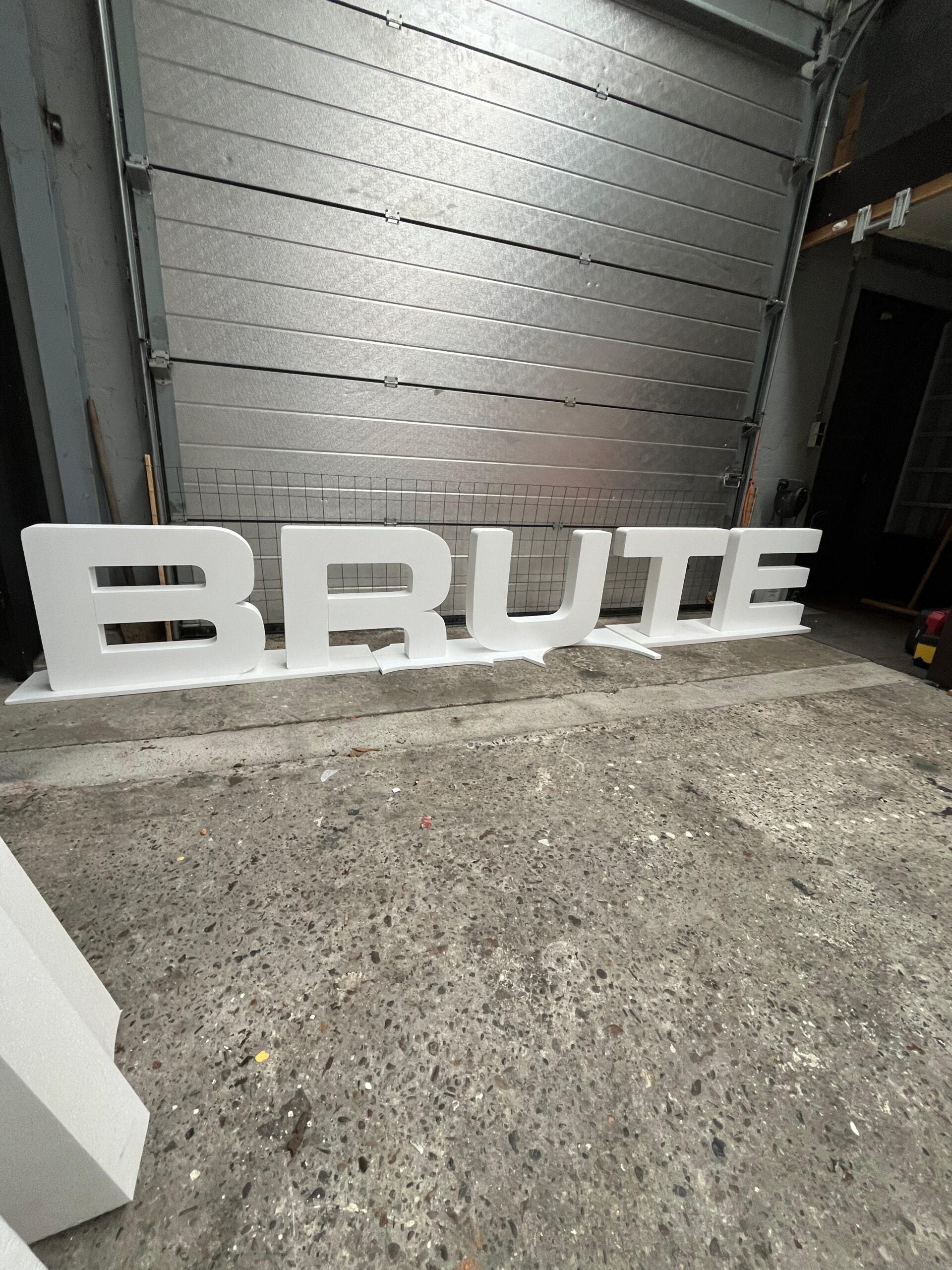 Large white letters spelling 'BRUTE' on a concrete floor with a metal garage door in the background.